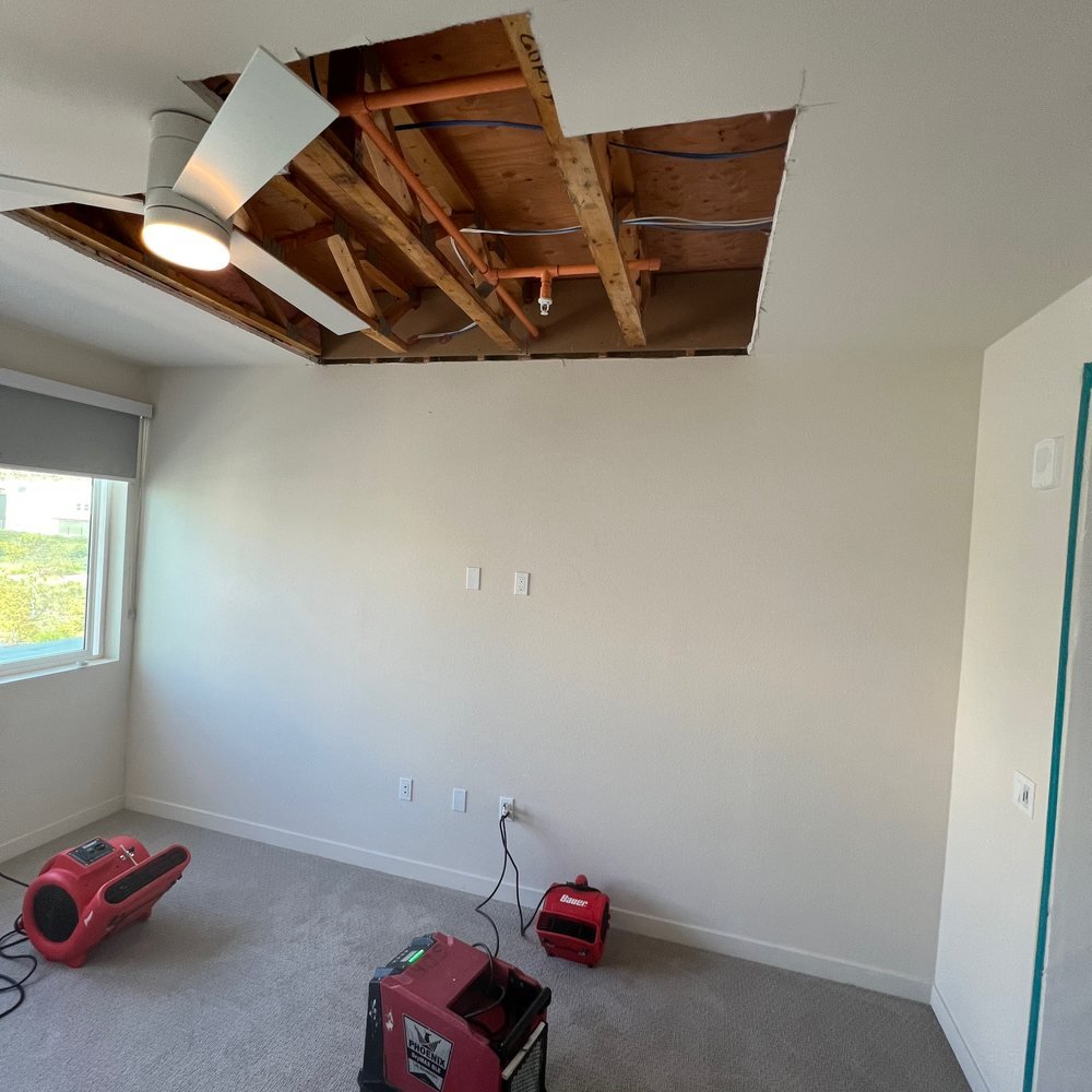 Bedroom ceiling opened to the joists after a water loss, with containment sheeting at the doorway and three air movers running on the carpet during structural drying